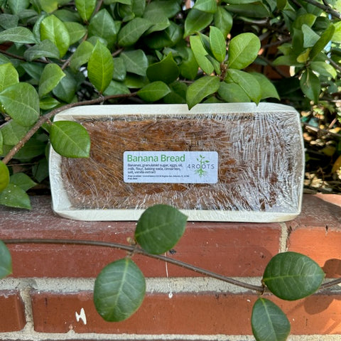 Banana bread in its package with a 4Roots label, standing atop a brick wall. The image highlights the product's attractive packaging and branding, showcasing the loaf’s freshness and quality. Ideal for emphasizing the product’s premium presentation and the 4Roots commitment to quality baked goods.
