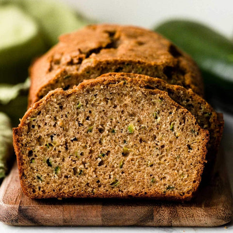 Close-up of a slice of zucchini bread showing bits of zucchini embedded in the moist loaf, with the rest of the loaf visible in the background. Freshly baked, the bread features a golden-brown crust and a tender, flavorful interior.